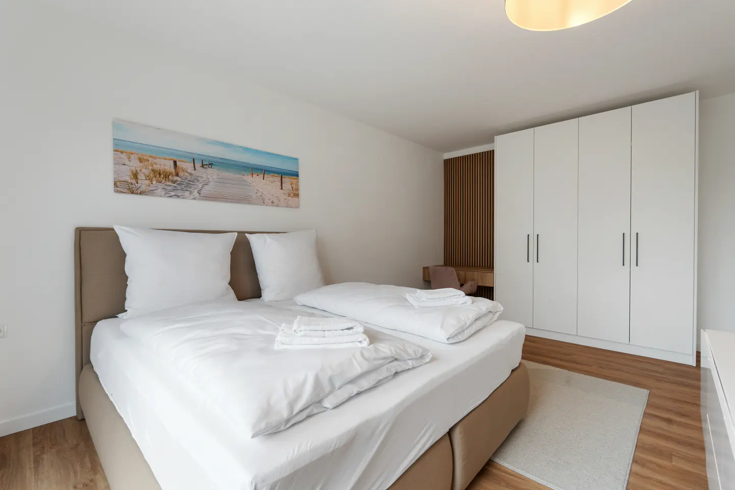 Bright bedroom with a tan bed frame, white linens, and a beach scene picture. A white wardrobe stands against the wall.
