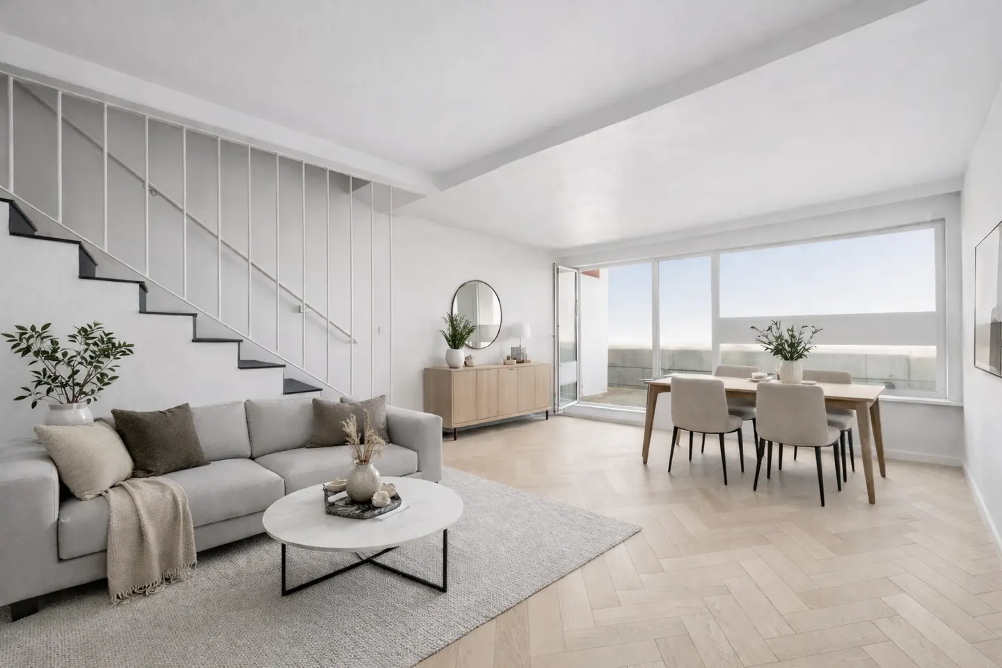 Bright, modern living room with a gray sofa, white coffee table, and dining set near a large window. Stairs are visible on the left.