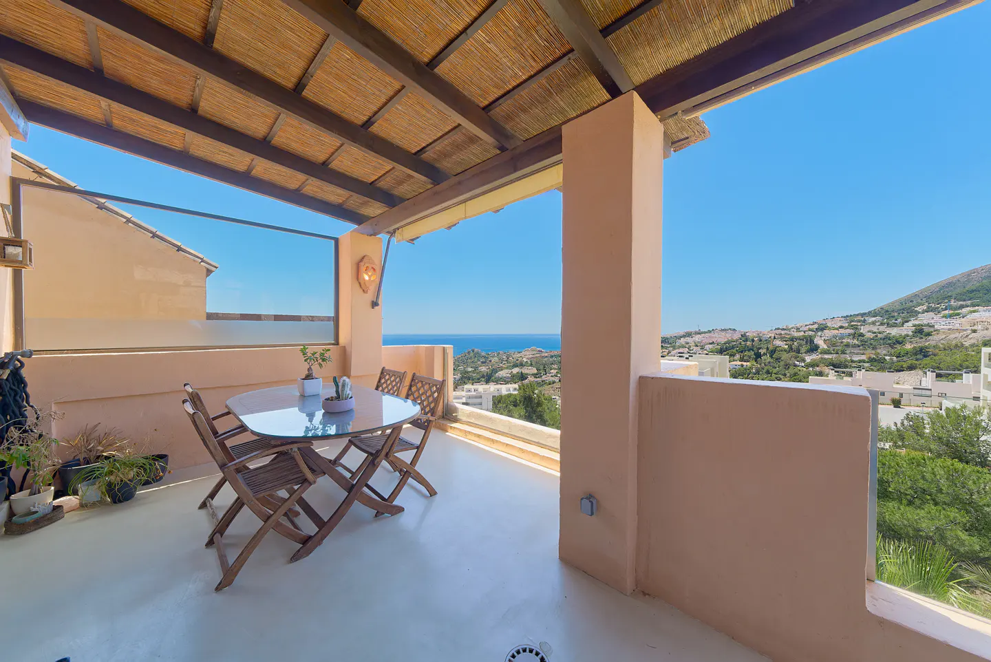 Outdoor patio with a glass table, four wooden chairs, and potted plants. The patio has a straw roof and overlooks the ocean and a hillside with white buildings.