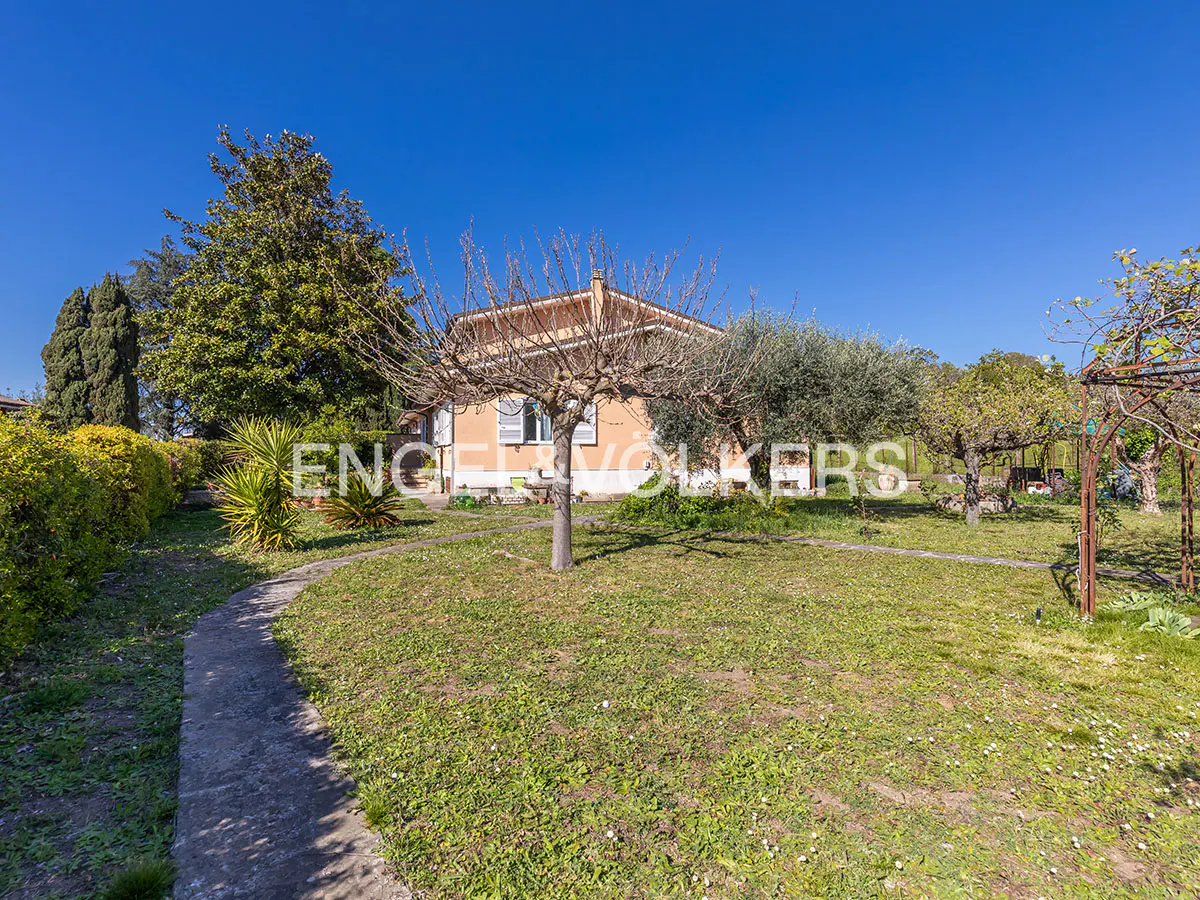 Exterior view of a single-story peach house with a large lawn, trees, and a stone path under a clear blue sky.