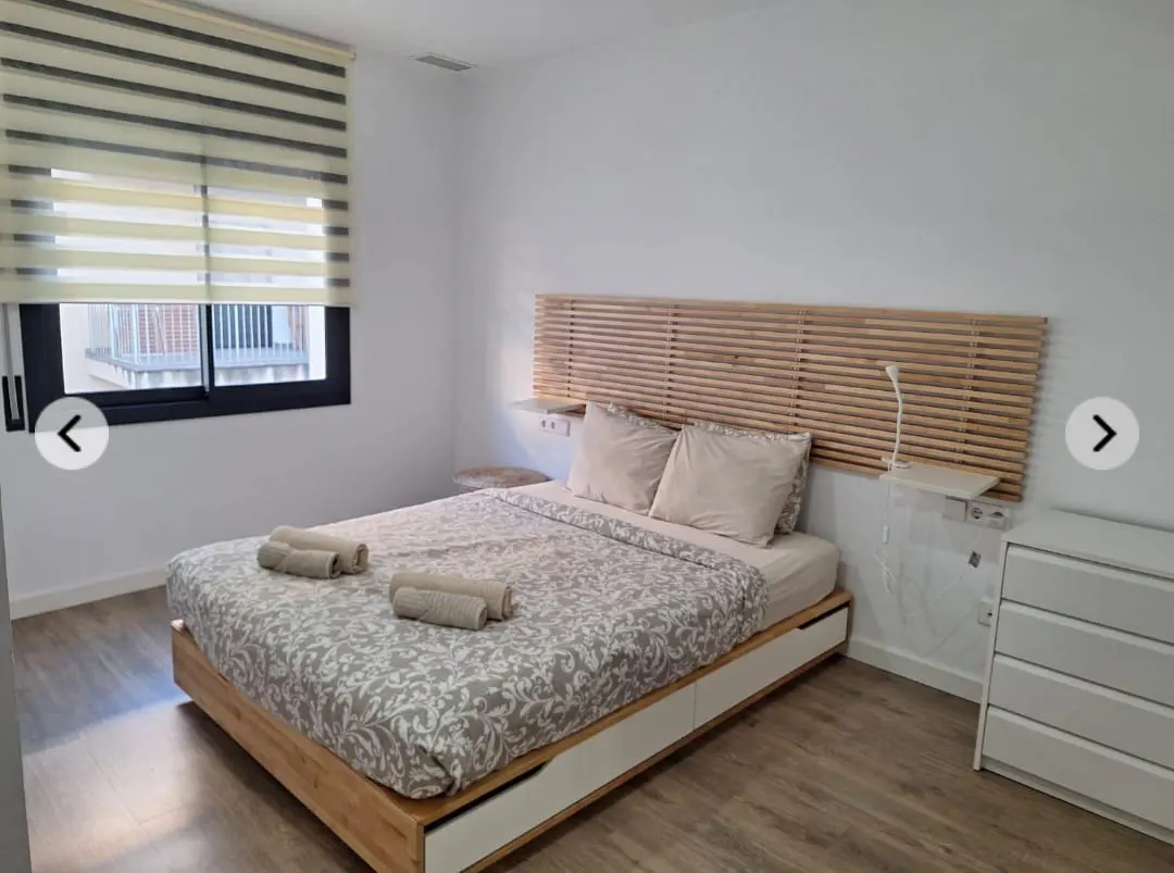 Bedroom with a wooden bed frame, patterned duvet, and rolled towels. A wooden headboard and white dresser are visible.