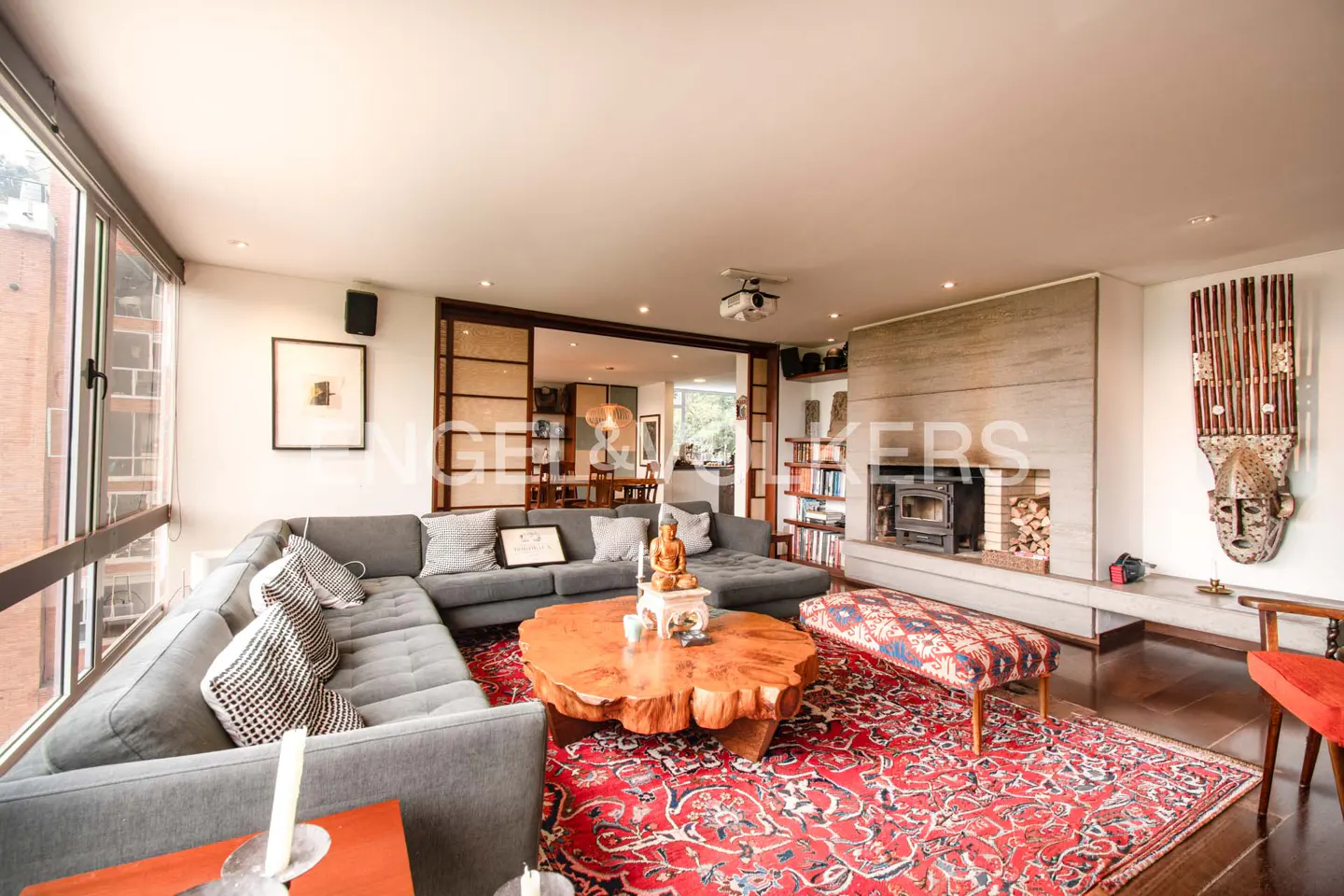 A living room with a gray sectional sofa, a wood coffee table, and a red patterned rug. A fireplace and a decorative mask are visible.