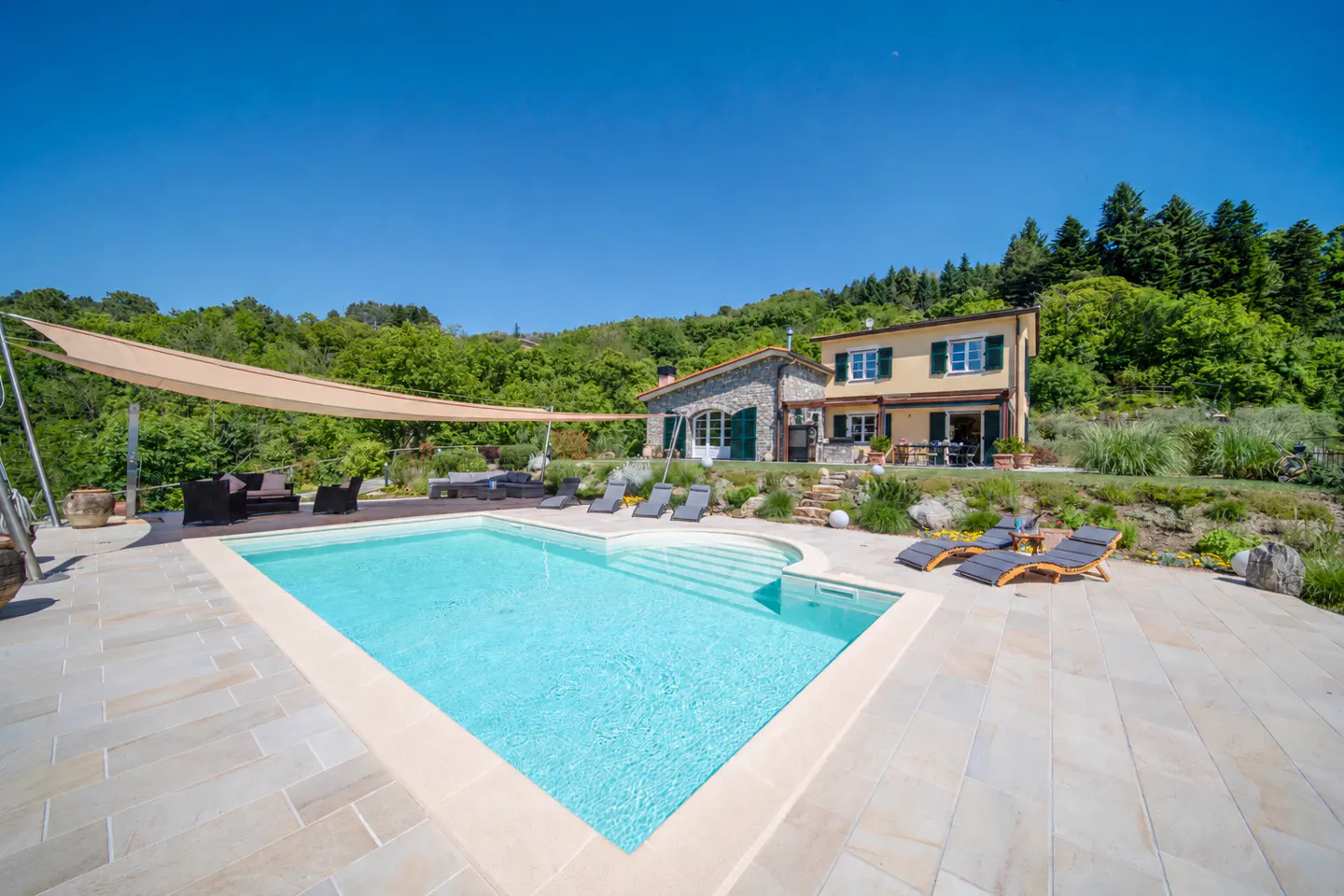 Exterior view of a two-story house with a pool, lounge chairs, and a tan sunshade. The house is yellow with green shutters and a stone facade.