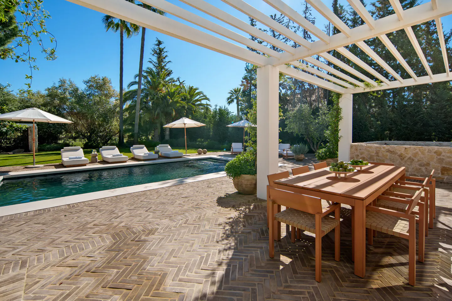 Outdoor patio with a wooden dining set under a white pergola, overlooking a swimming pool with lounge chairs and palm trees.