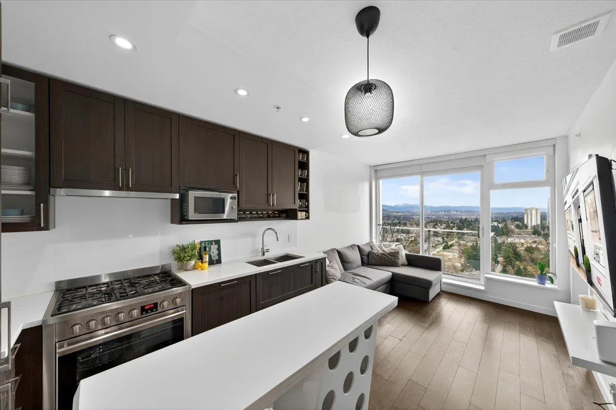 A modern condo kitchen with dark wood cabinets, stainless steel appliances, and a white countertop island. A gray sofa sits near a large window with a city view.