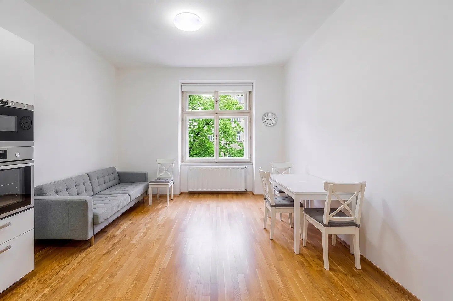 Bright, airy apartment with hardwood floors, white walls, and a large window. A gray sofa sits near a modern oven, and a white dining set is by the window.