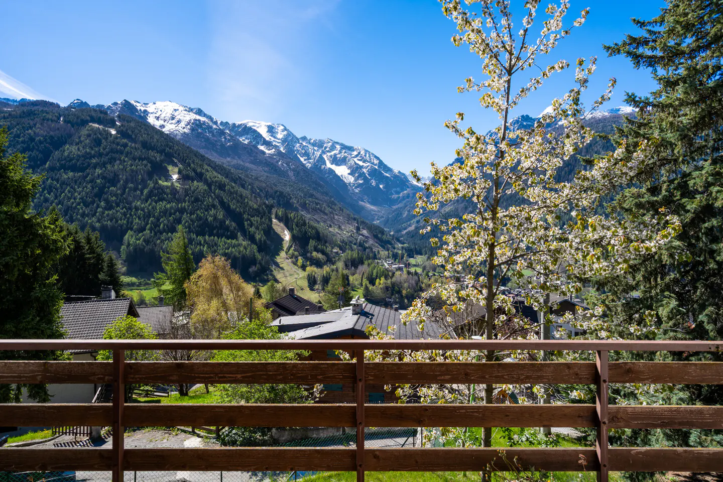 View from a wooden balcony overlooking a valley with snow-capped mountains, green trees, and houses under a blue sky.