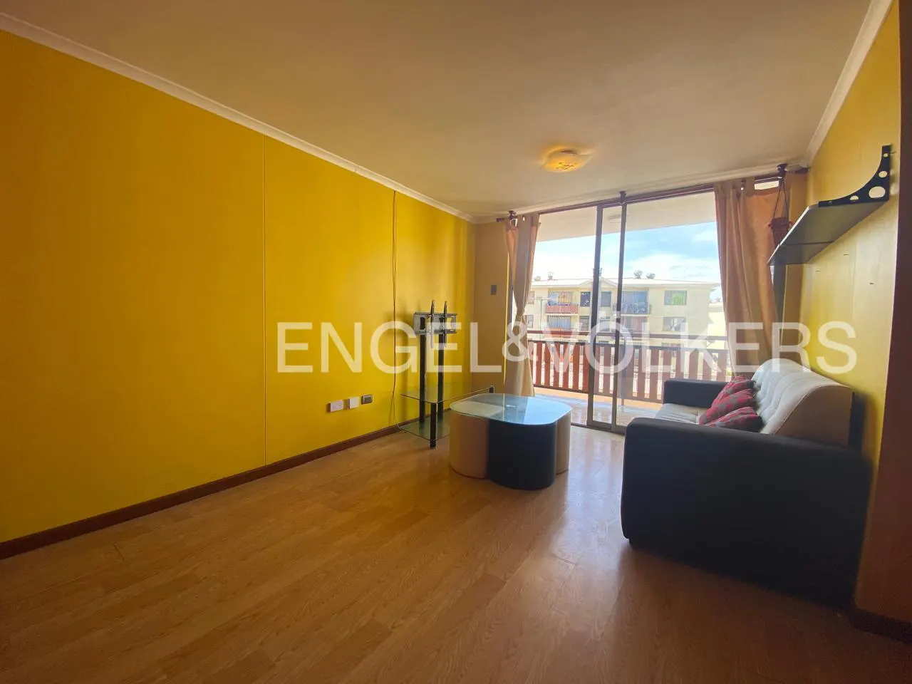 Bright living room with yellow walls, wood floors, and a black sofa. Sliding glass doors lead to a balcony. A modern coffee table sits in the center.