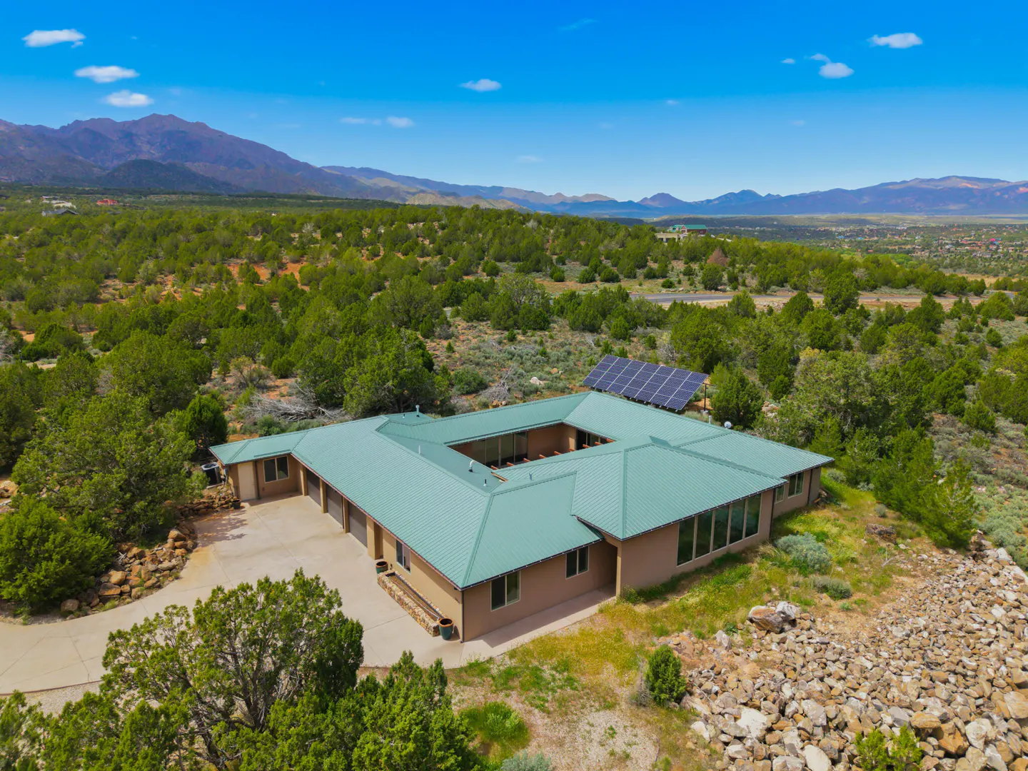 Aerial view of a U-shaped, tan house with a green metal roof and solar panels, surrounded by green trees and mountains.