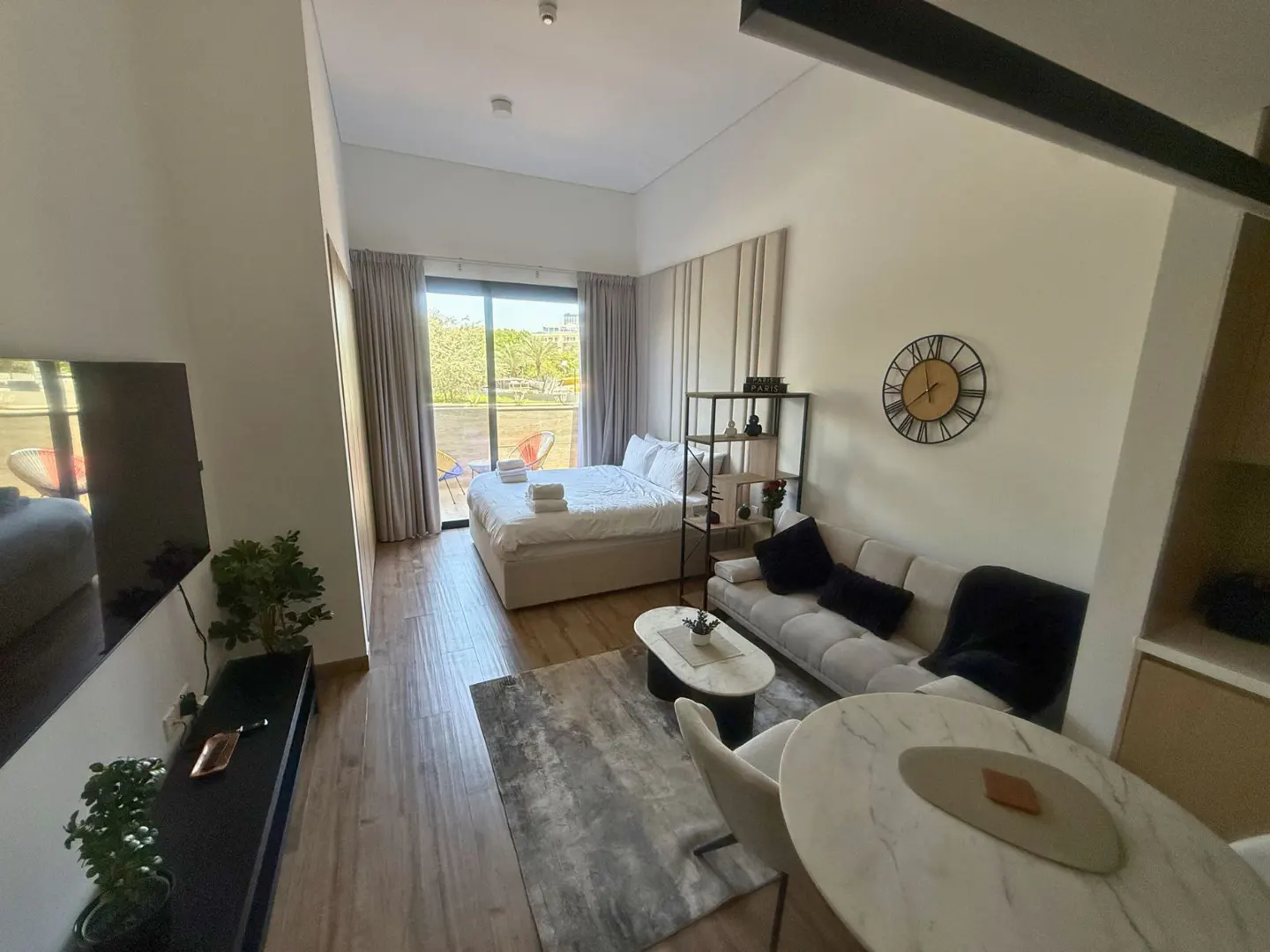 Studio apartment with a bed, sofa, marble table, and TV. A balcony overlooks a green space. The interior is decorated in neutral tones.