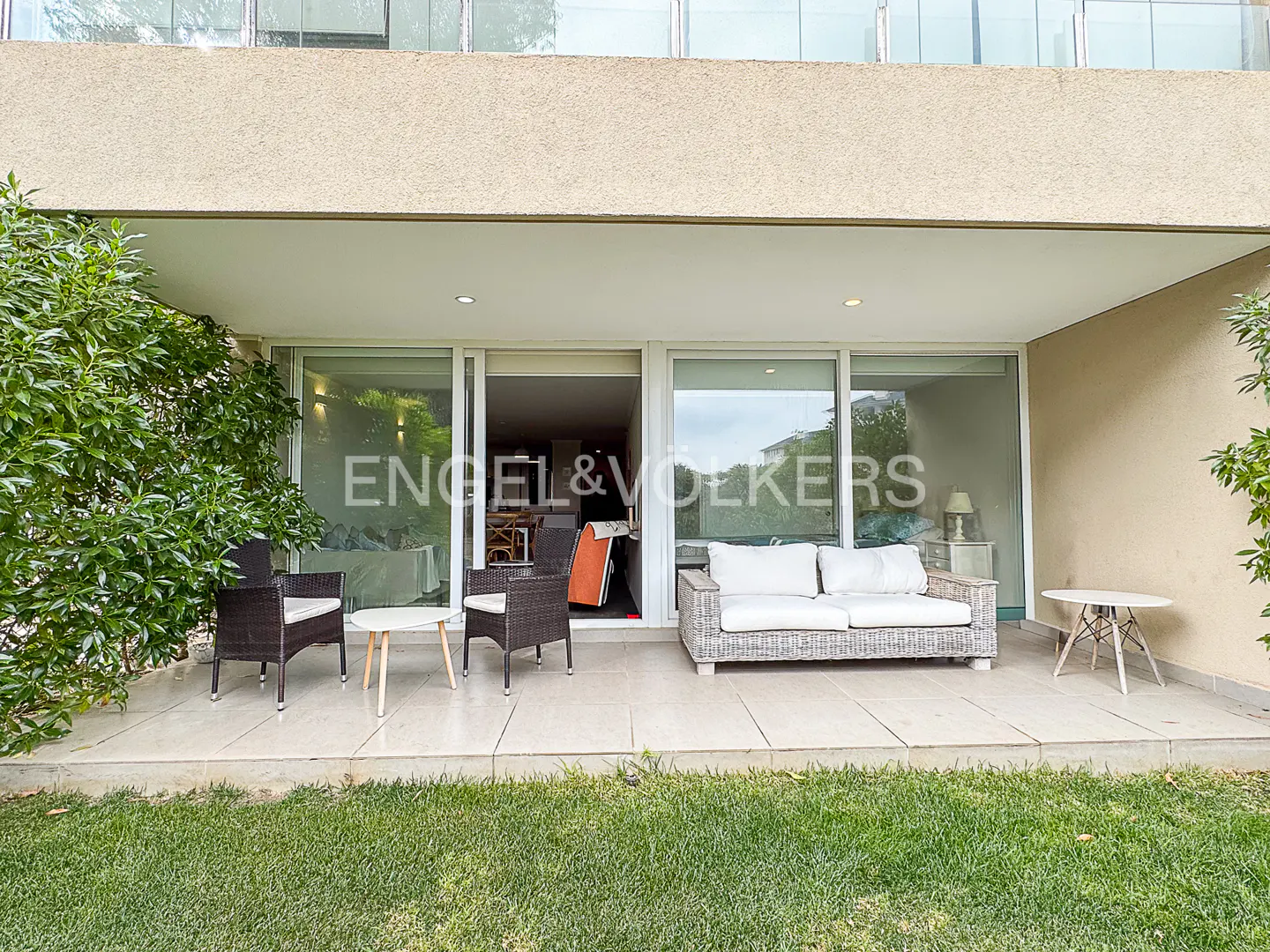 Covered patio with wicker furniture, white cushions, and small tables, overlooking a green lawn outside a modern home.