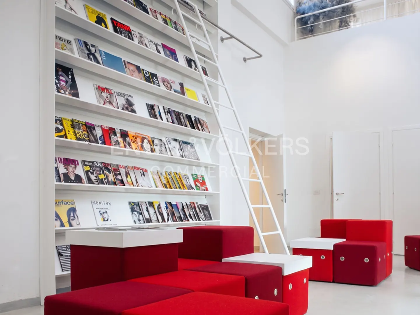 Bright, modern office space with a white bookshelf filled with magazines and a white ladder. Red modular seating is in the foreground.