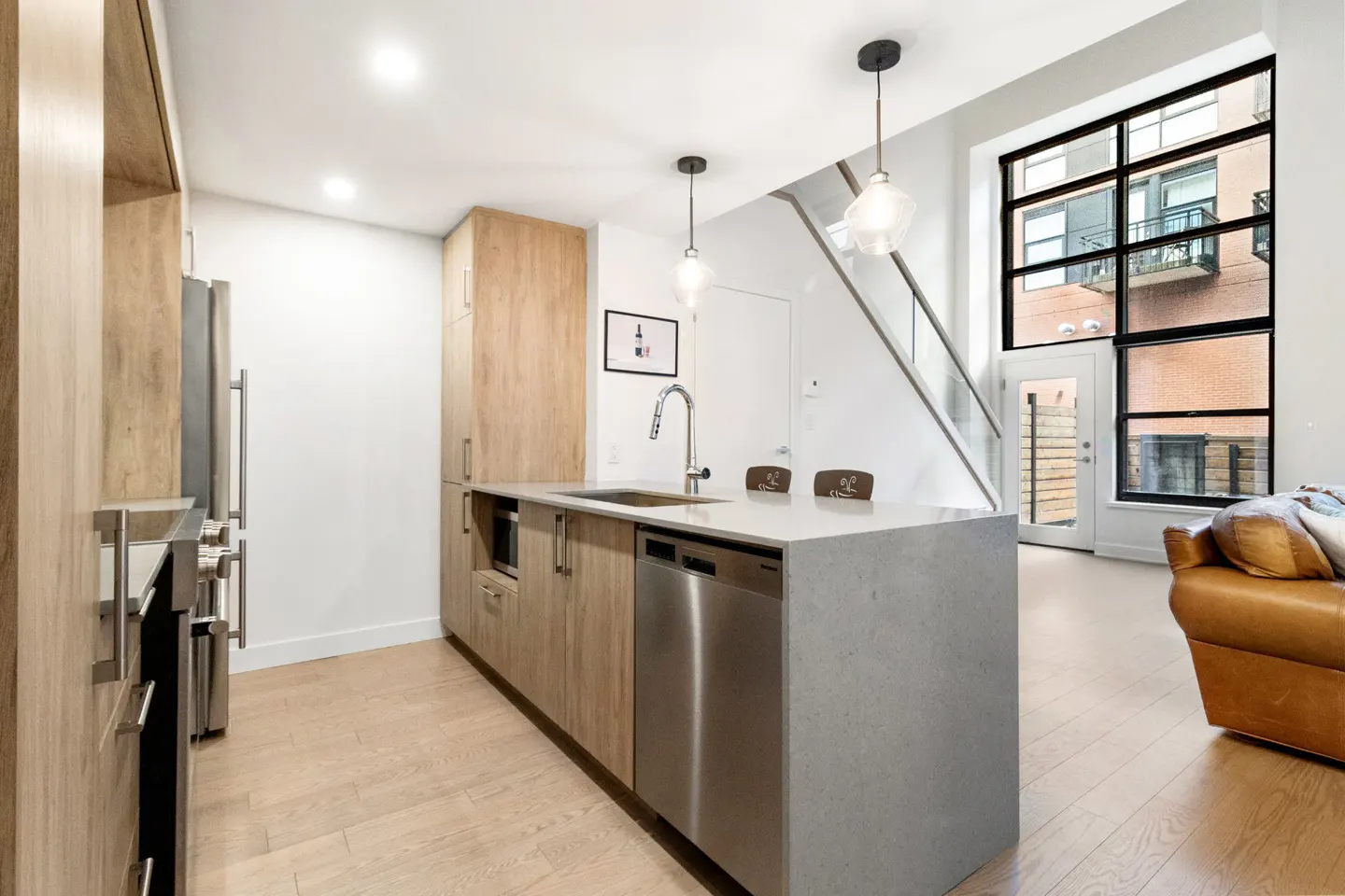 Modern kitchen with wood cabinets, stainless steel appliances, and a gray island. A large window and staircase are visible in the background.
