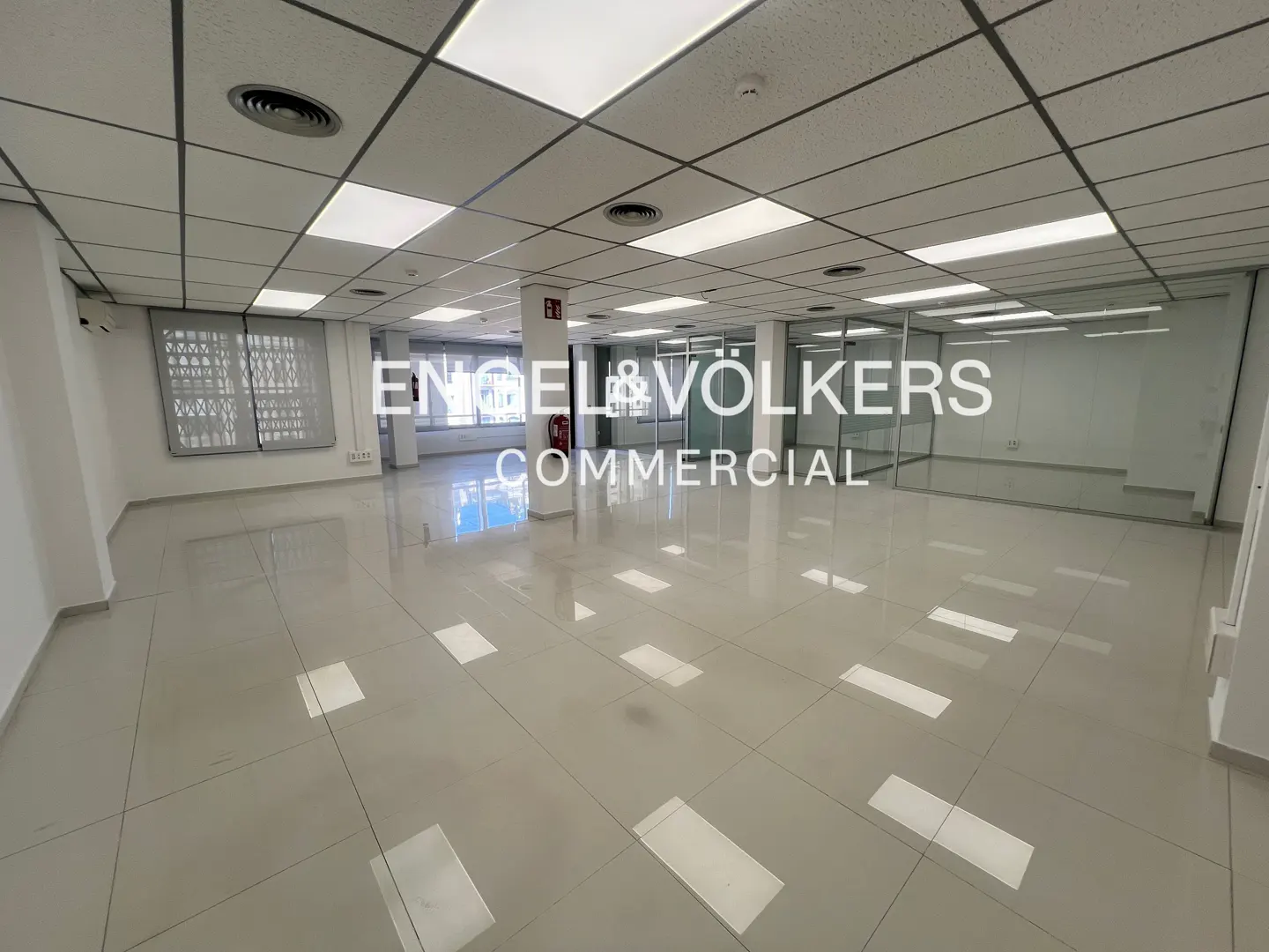 Bright, empty commercial space with white walls, tile floors, and a drop ceiling with recessed lighting. Glass-walled offices are visible in the background.