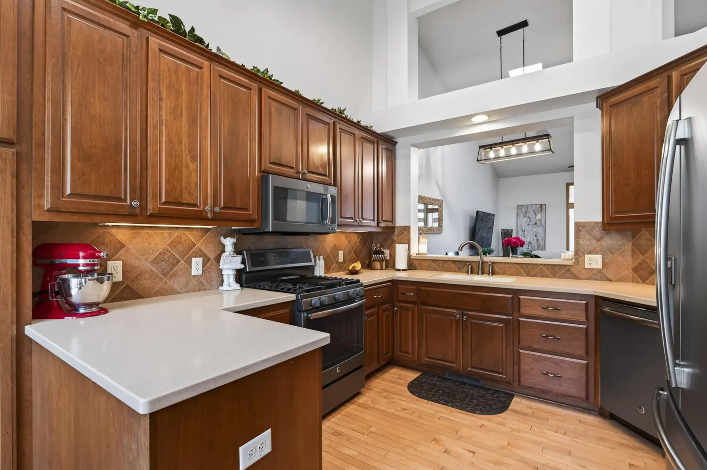 A kitchen with wood cabinets, stainless steel appliances, and a white countertop. A red stand mixer sits on the counter. A pass-through looks into the living room.
