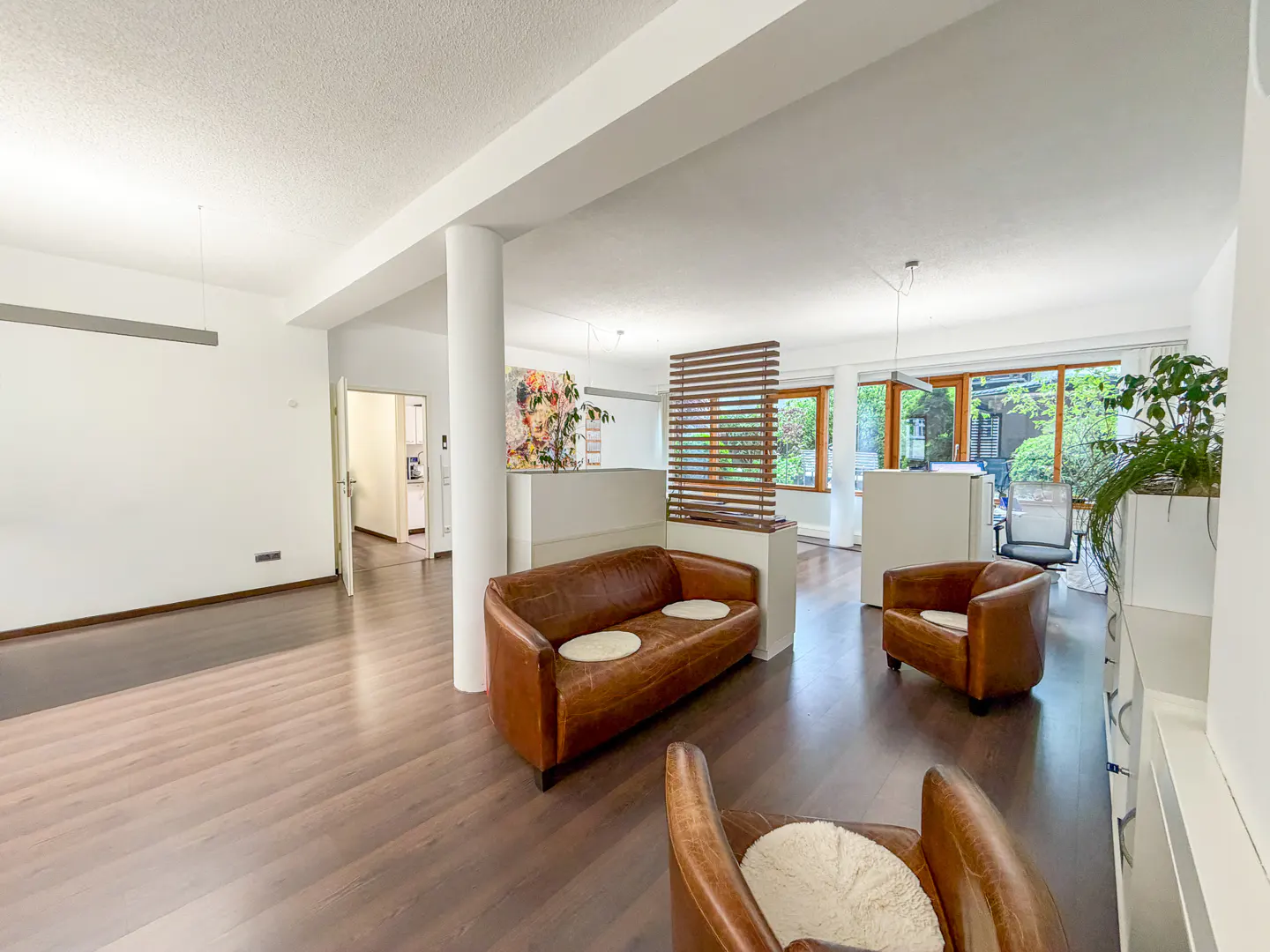 A bright living room with brown leather sofas and chairs on a wood floor. White walls and pillars divide the space. Windows overlook greenery.