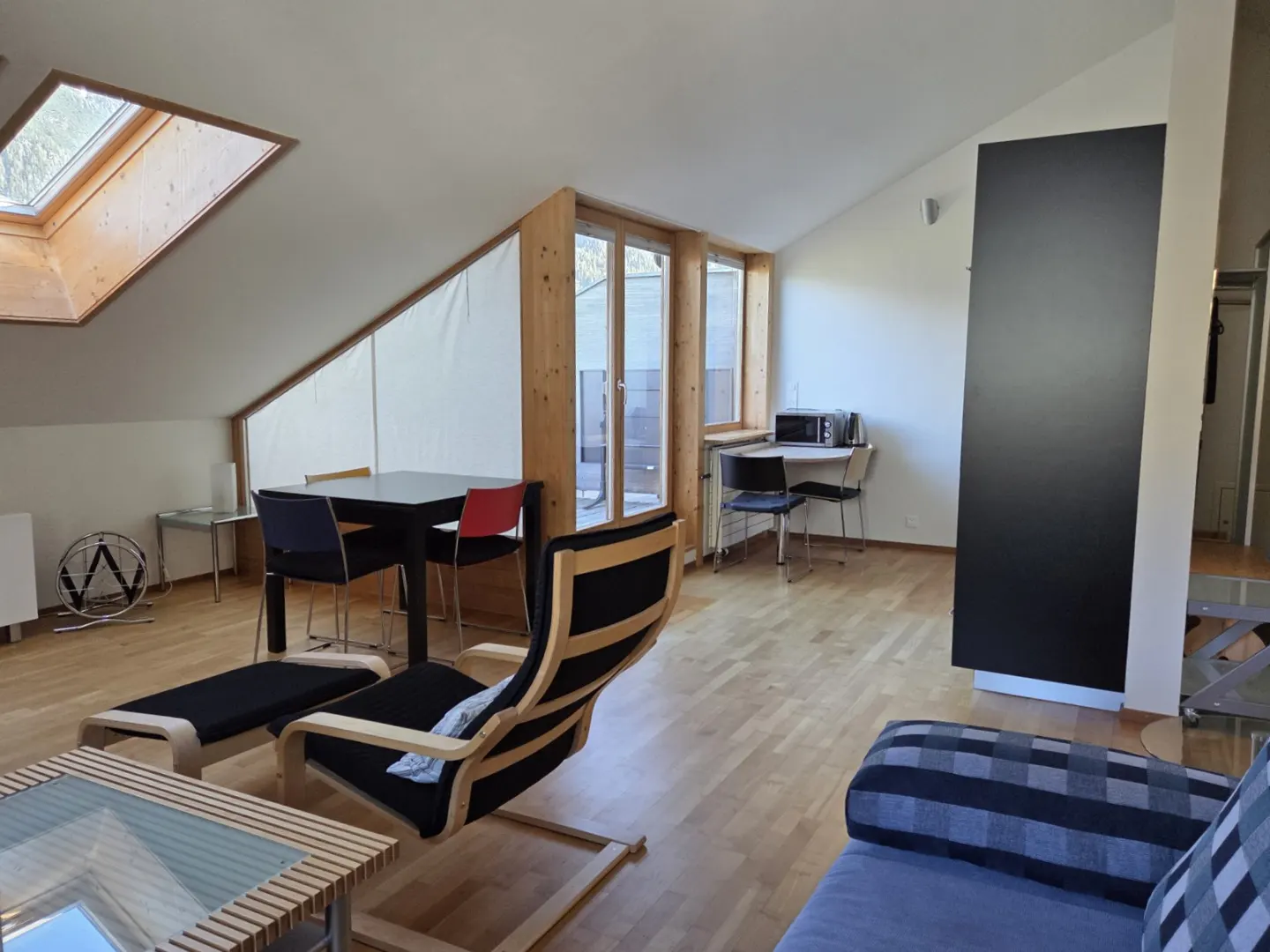 Attic apartment interior with wood floors, a black dining table with blue and red chairs, and a black lounge chair with ottoman.