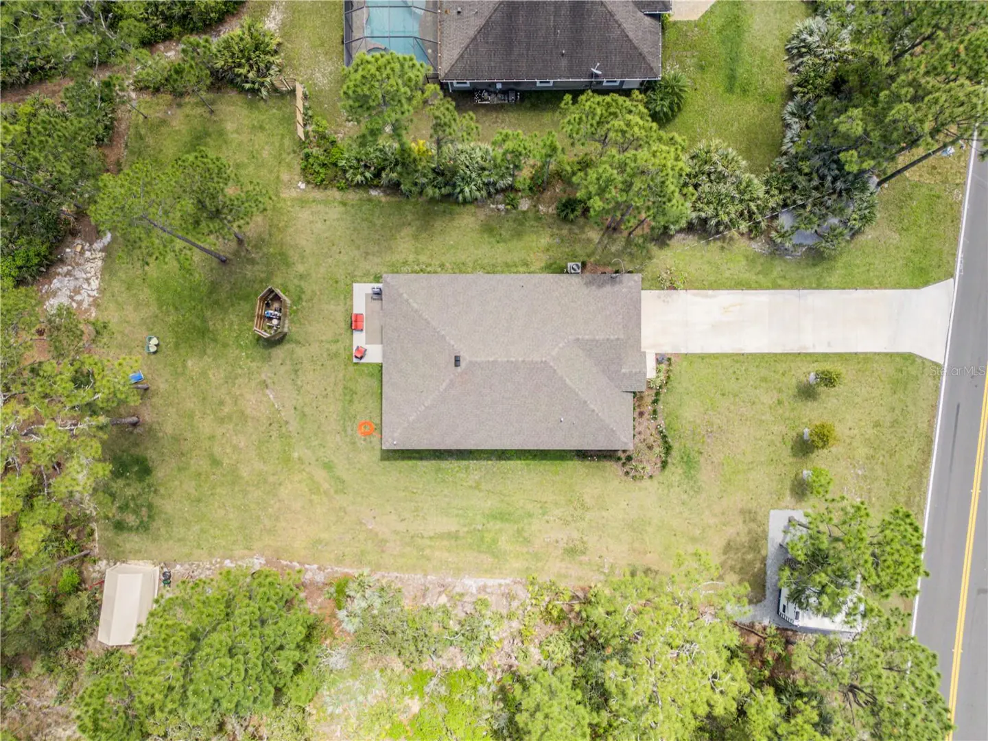 Aerial view of a single-story home with a gray roof, concrete driveway, and large grassy yard surrounded by pine trees.