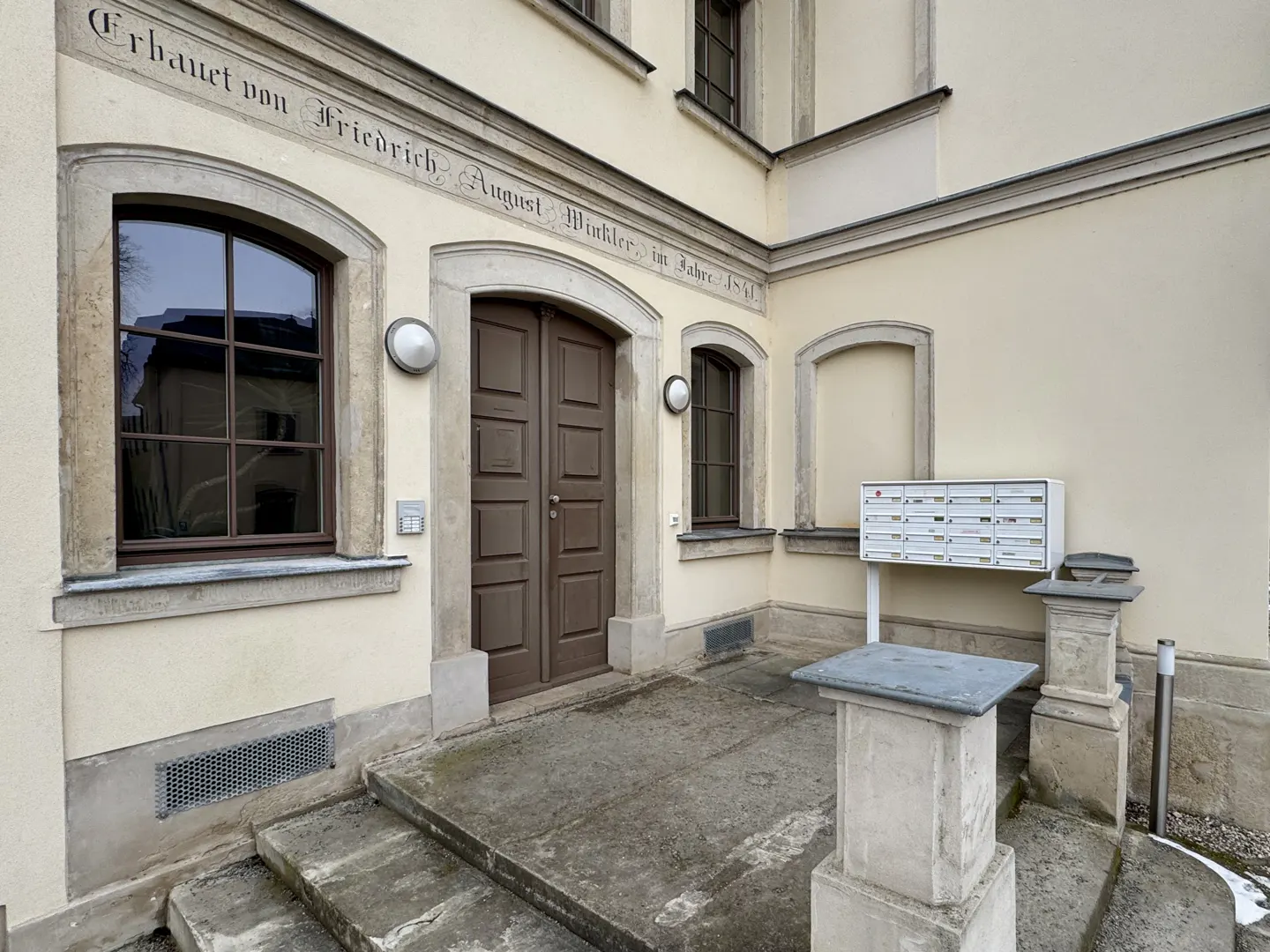 Exterior view of a cream-colored building with a brown door, windows, and a white mailbox. Inscription above the door.