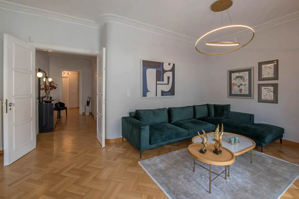 A modern living room with a green velvet sectional sofa, wood floors, and white walls. A gold ring chandelier hangs above a coffee table with gold bird sculptures.