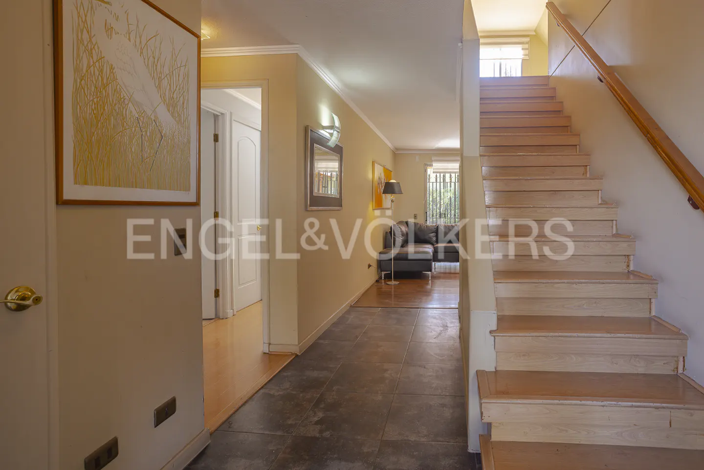 A home's interior view shows a staircase, hallway, and living room. Artwork hangs on the walls. The living room has a dark sofa.