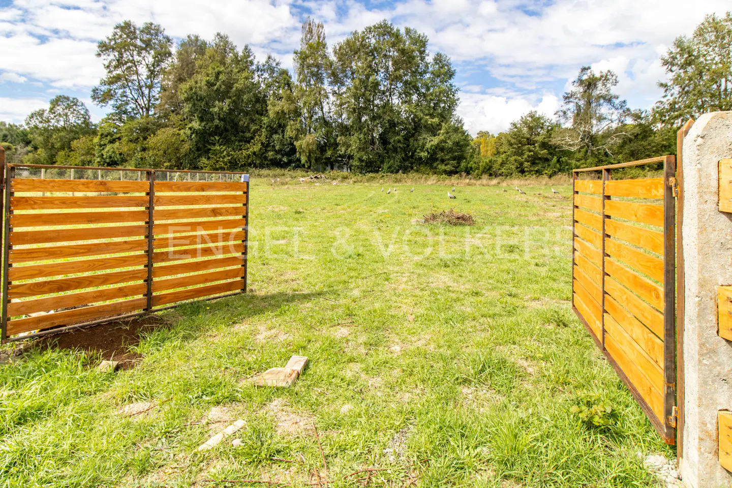 Open wooden gate leads to a grassy field with trees in the background under a partly cloudy sky.