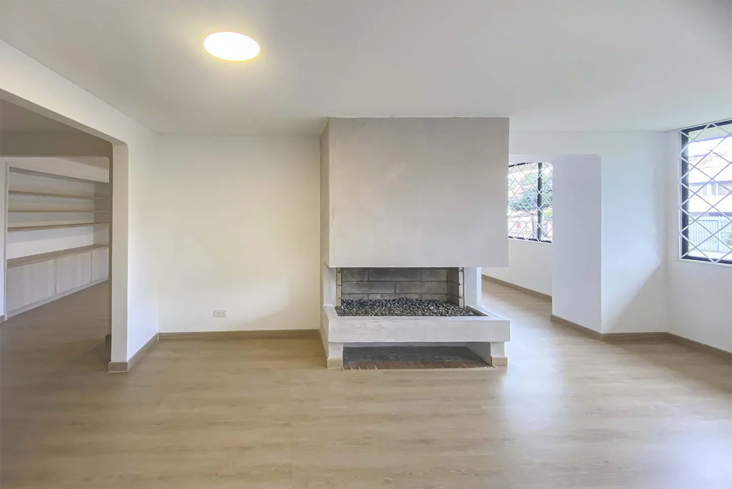 Bright, empty living room with light wood floors, white walls, and a modern fireplace with gray stones. Windows let in natural light.