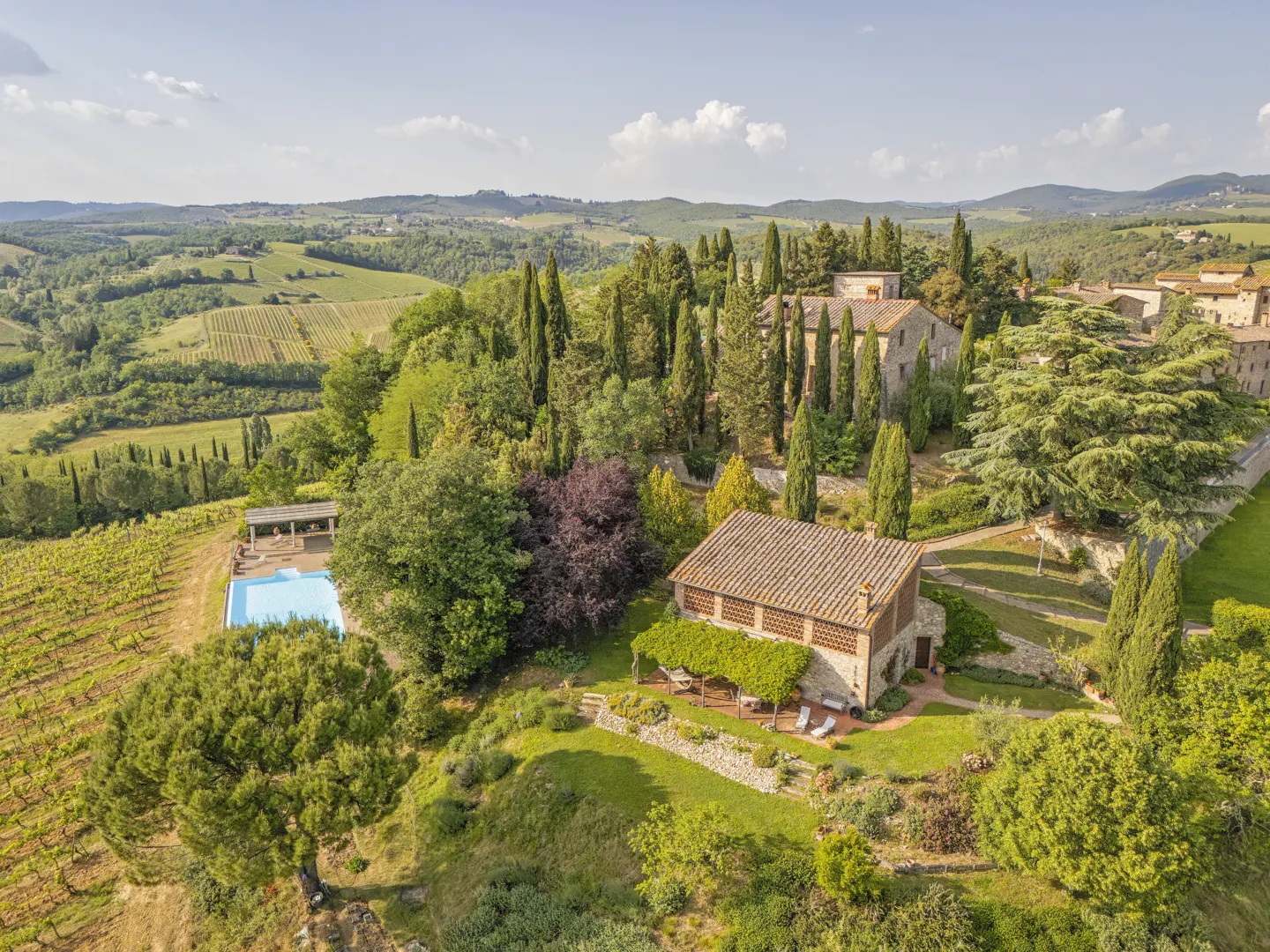 Aerial view of a Tuscan villa with a pool, surrounded by lush gardens, cypress trees, and rolling vineyard hills.