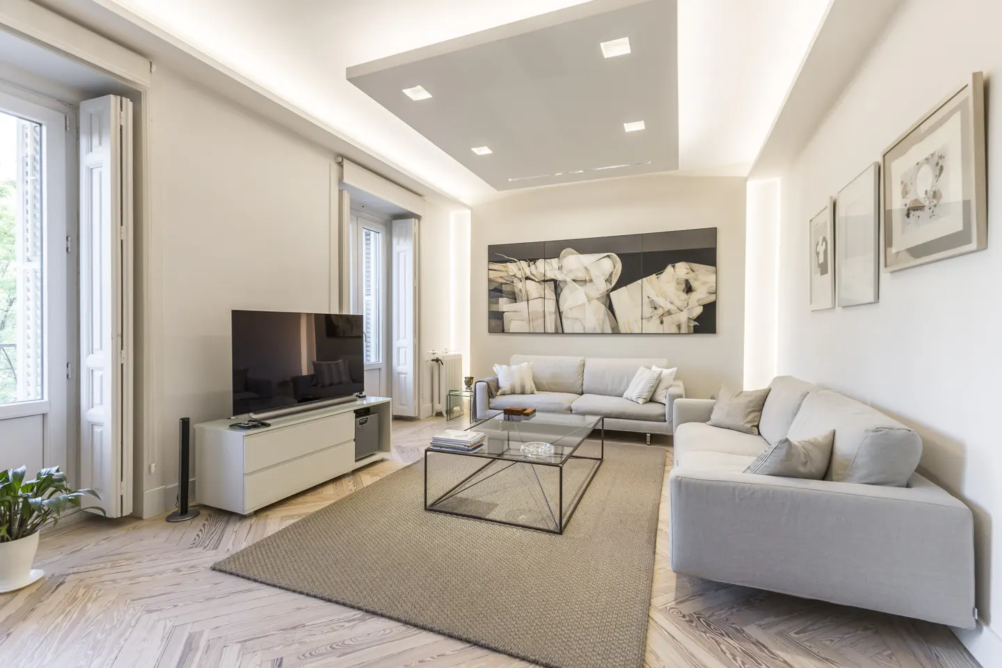 Bright living room with light gray sofas, a glass coffee table on a tan rug, and a TV on a white console. Art adorns the white walls.
