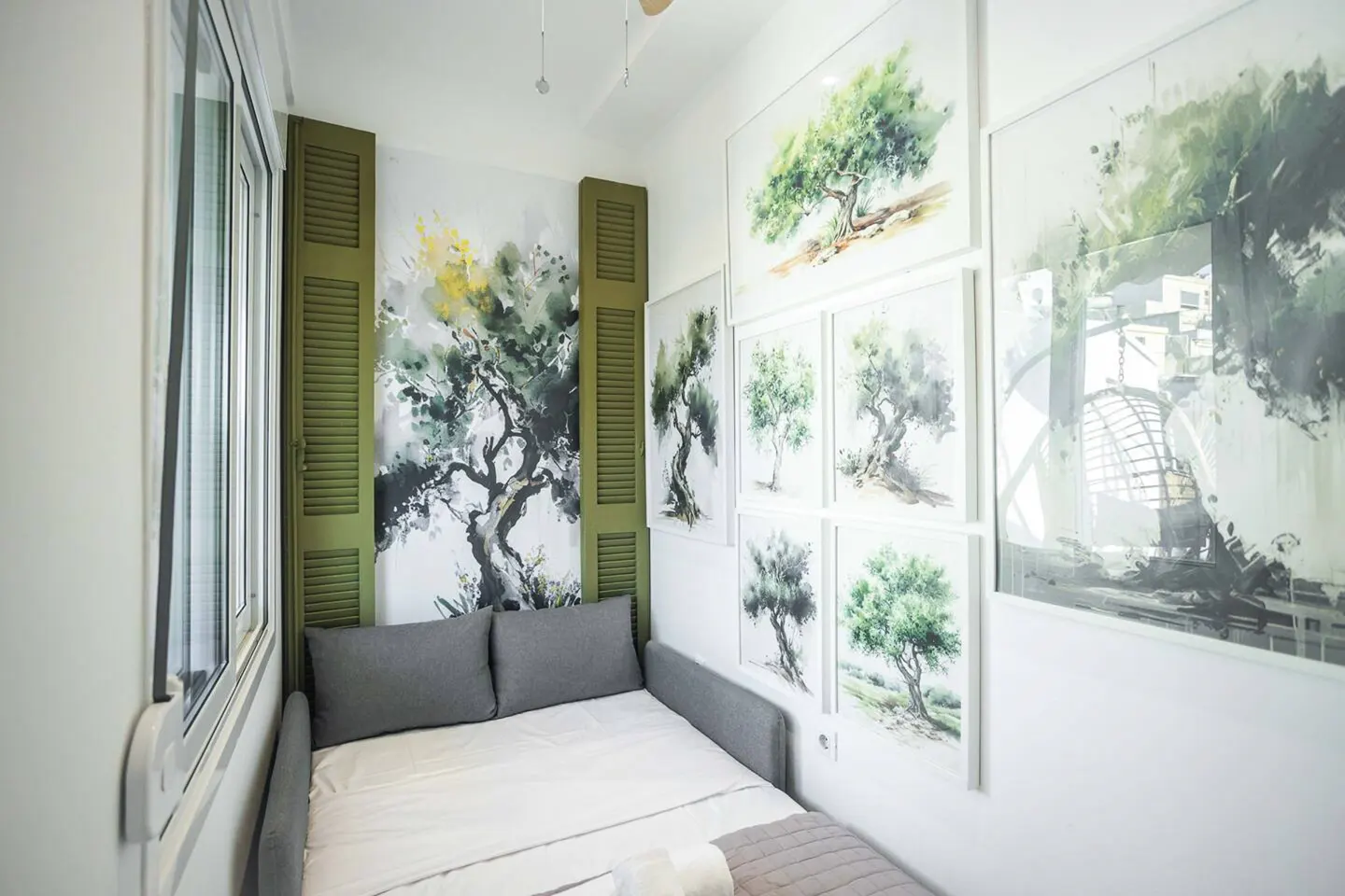 A small bedroom with a gray sofa bed, white walls, and tree-themed artwork. Green shutters flank a tree mural.
