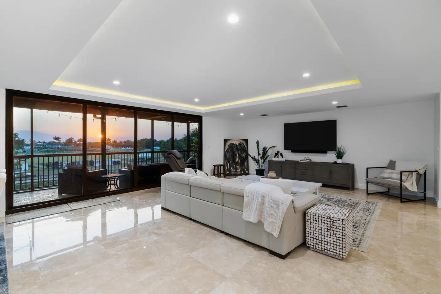 Bright living room with a beige sectional sofa, marble floors, and a sunset view from the balcony. A large TV is mounted on the wall.