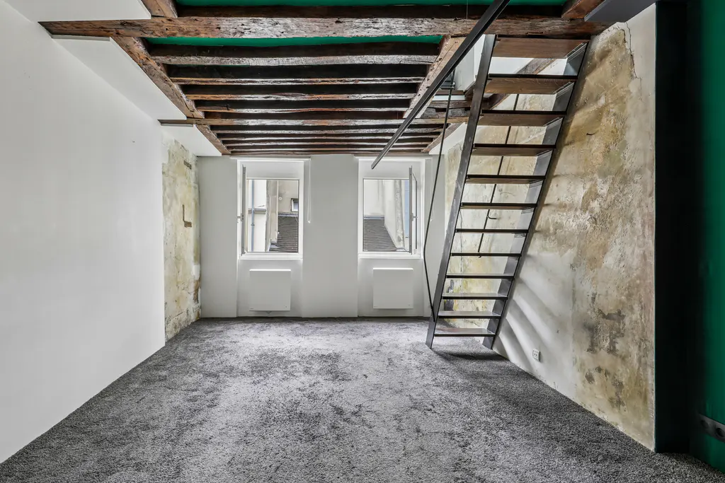 Empty room with gray carpet, exposed wooden ceiling beams, metal staircase, and two windows overlooking a courtyard.