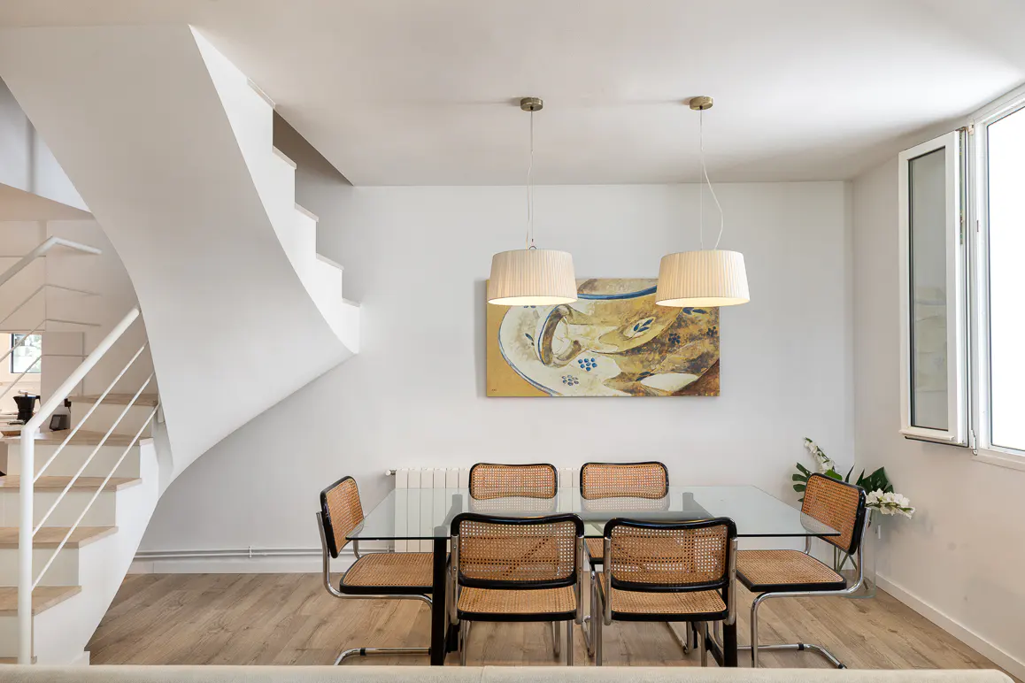 Modern dining area with a glass table, six cane-back chairs, two pendant lights, and a staircase in a bright, white room.