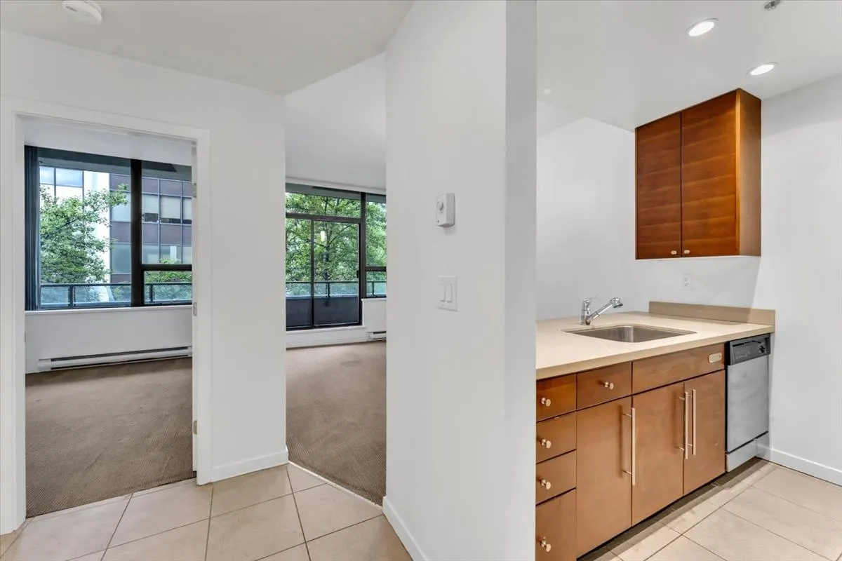 A bright apartment interior shows a kitchen with wood cabinets, a sink, and a dishwasher. A doorway leads to a living room with large windows.