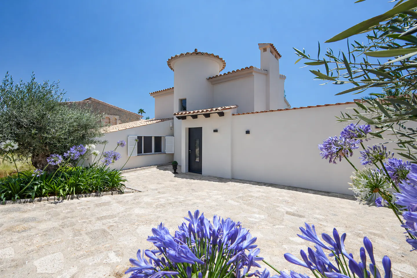 Exterior view of a white stucco house with a turret, a black door, and a stone driveway, framed by blue agapanthus flowers.