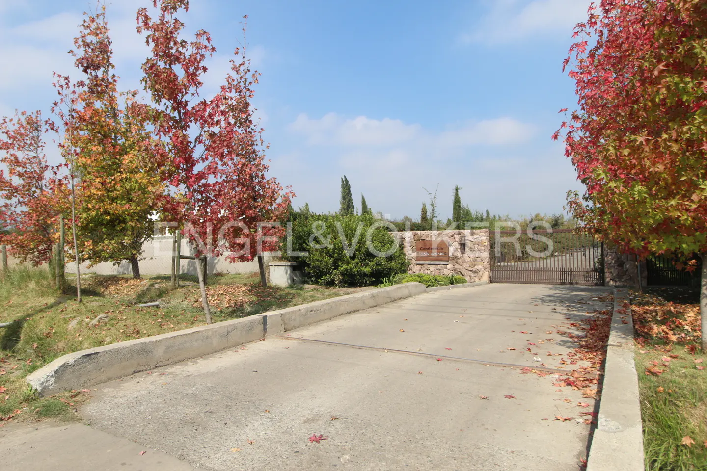 Gated driveway entrance to a property with red-leaved trees and a stone wall under a blue sky.