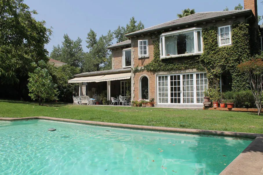 Exterior view of a brick house with a pool, lawn, and patio furniture on a sunny day.