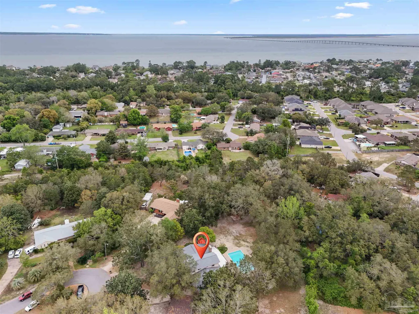 Aerial view of a house with a pool, surrounded by trees, with a red location pin on the roof. A bay and bridge are in the background.