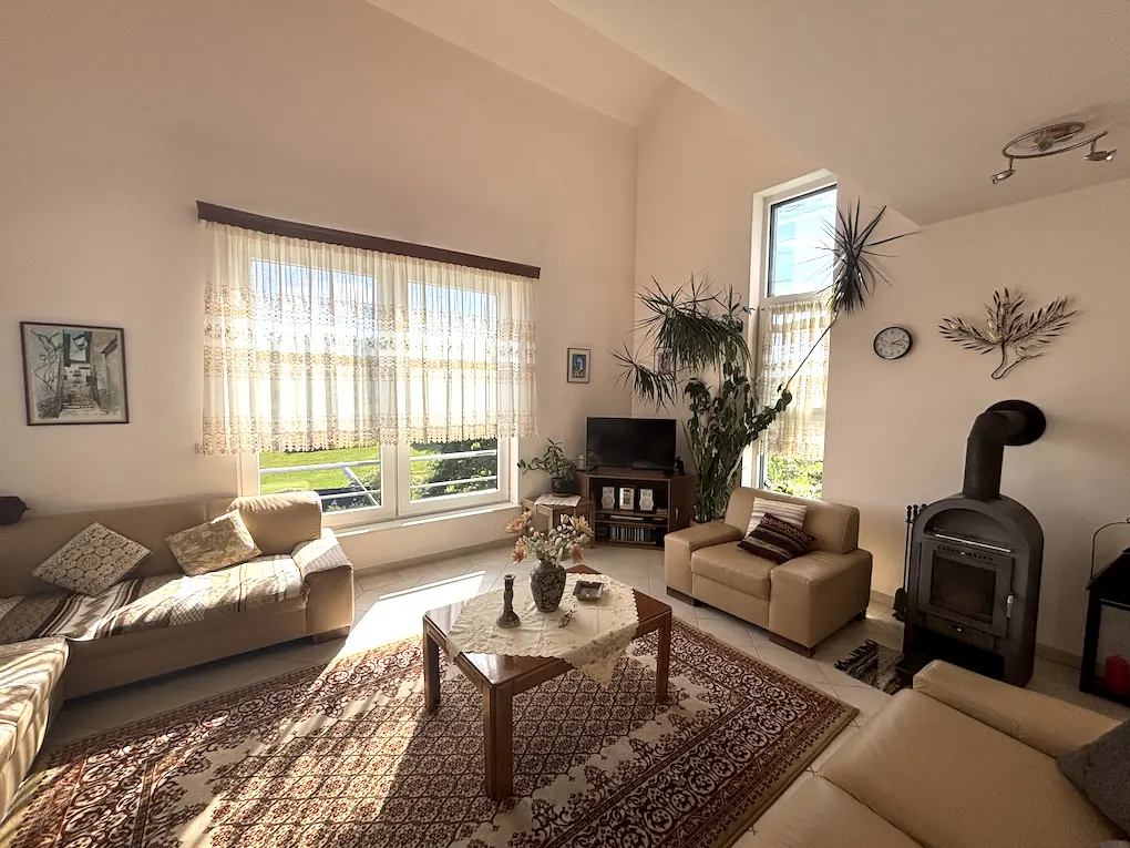 Living room with beige sofas, a wood-burning stove, and a large window with sheer curtains. A patterned rug covers the floor.