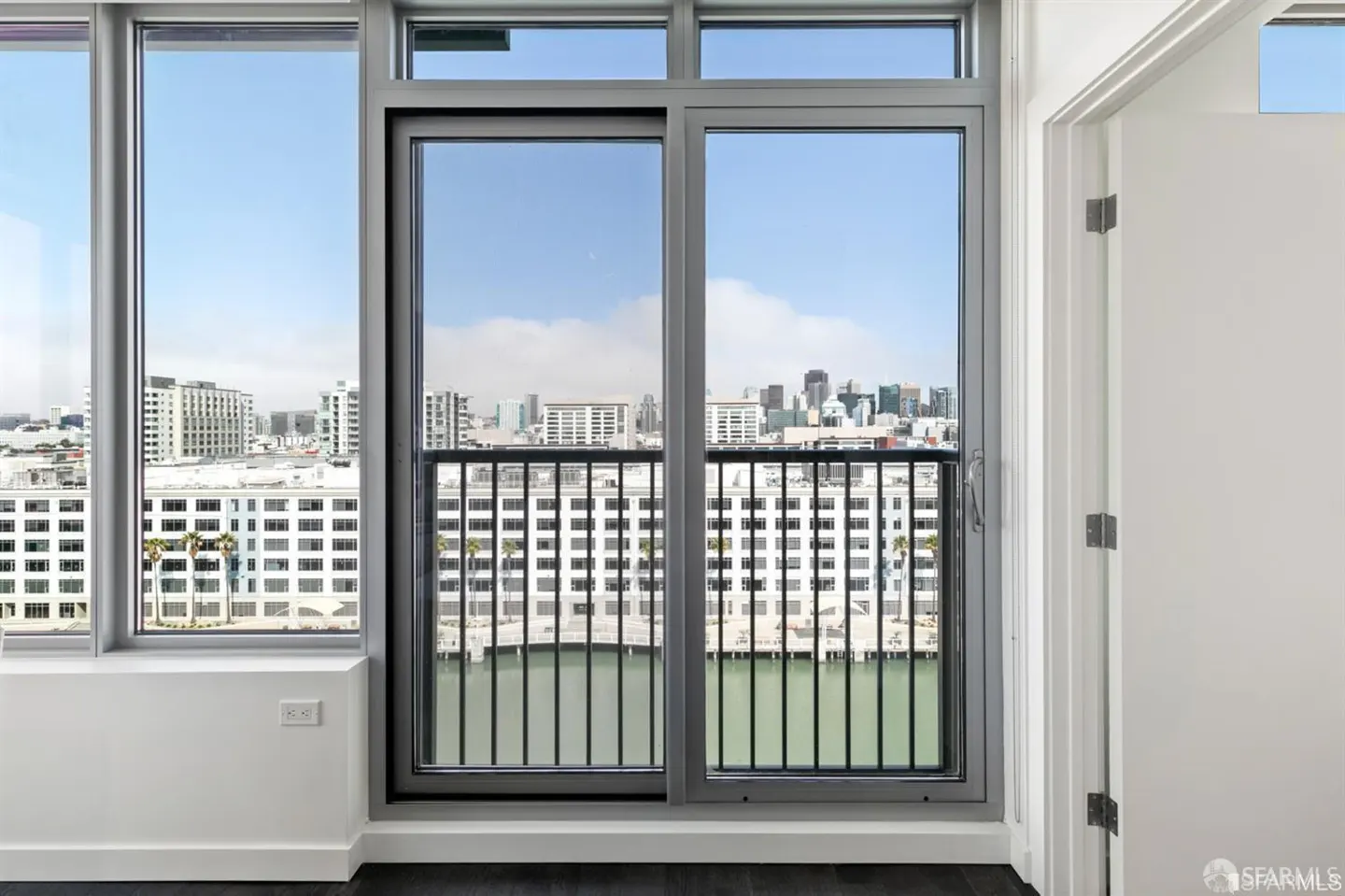 View from a condo with a sliding glass door to a balcony overlooking a city skyline and bay. A white door is partially visible on the right.