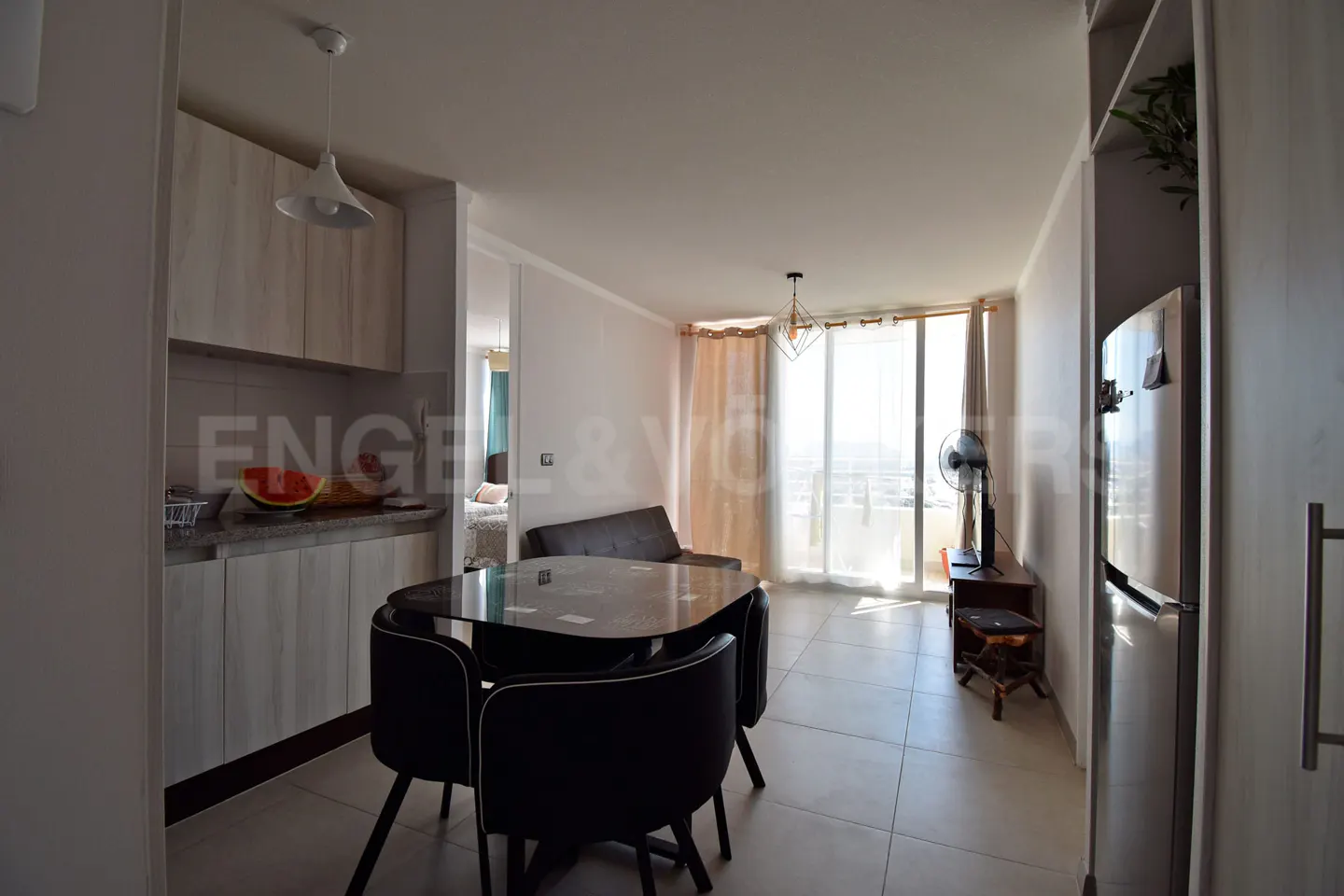 Bright apartment interior with kitchen cabinets, black table and chairs, sofa, and balcony with sheer curtains.