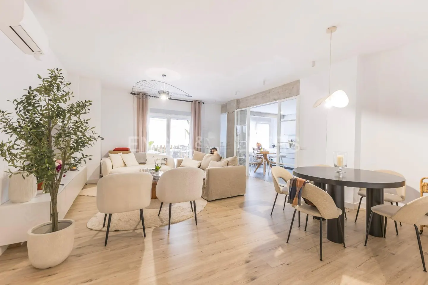 Bright, open-plan living and dining area with light wood floors, neutral furniture, and a large potted plant. Glass doors lead to another room.