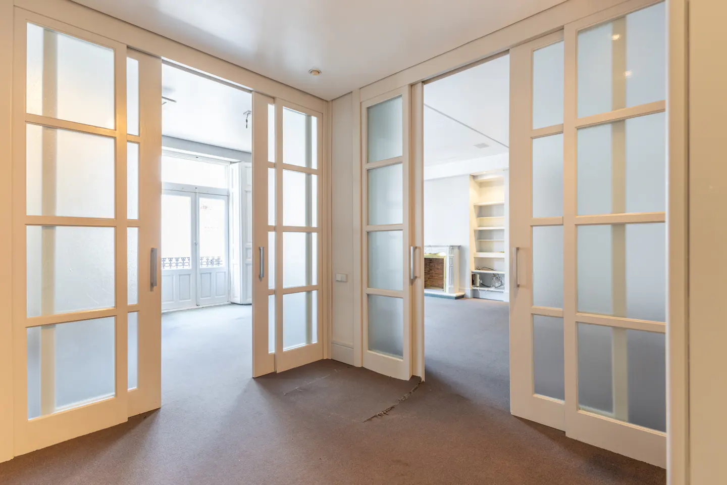 Interior view of a room with sliding white doors, frosted glass panes, and a gray carpet. A fireplace and shelves are visible in the adjacent room.