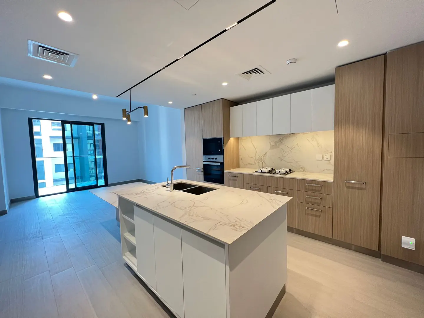 A modern kitchen with white marble countertops, a central island, and light wood cabinets. Sliding glass doors lead to a balcony.