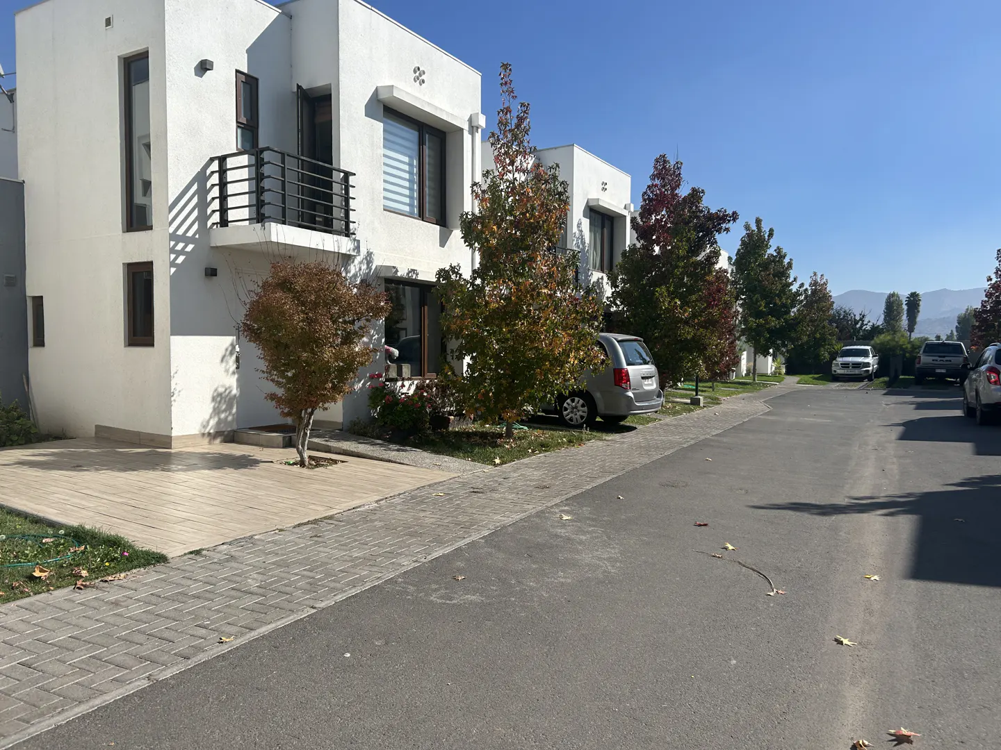 Street view of modern white townhouses with black balconies, trees, and parked cars under a clear blue sky.