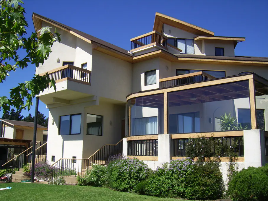 A multi-story beige house with balconies and a glass-enclosed patio under a blue sky. Green lawn and bushes in the foreground.