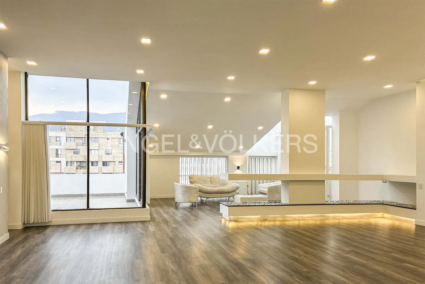 Bright, modern living room with wood floors, white walls, and large windows with city views. Seating area with white furniture.