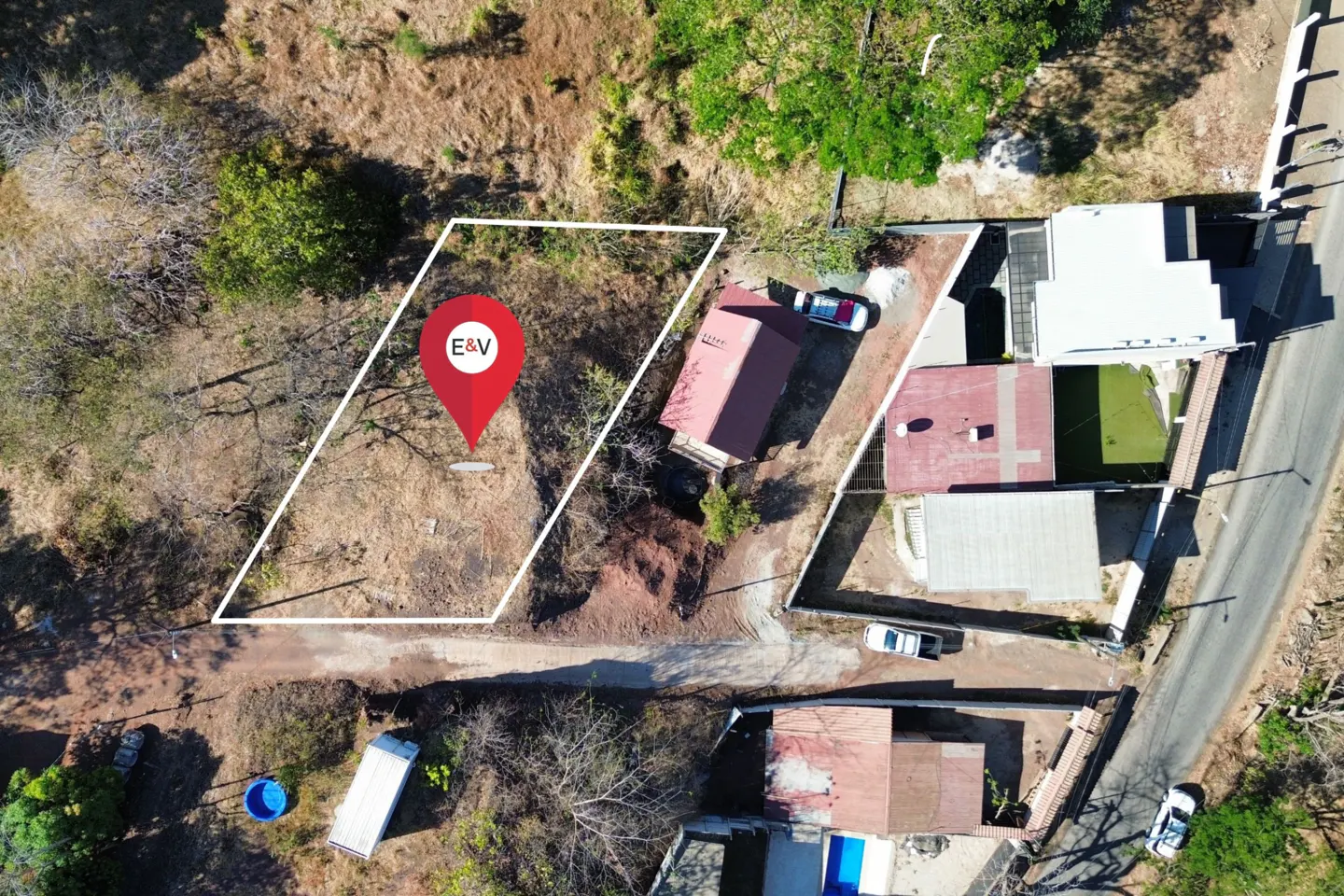 Aerial view of a vacant lot outlined in white, with a red pin marker labeled "E&V". Houses and trees surround the property.