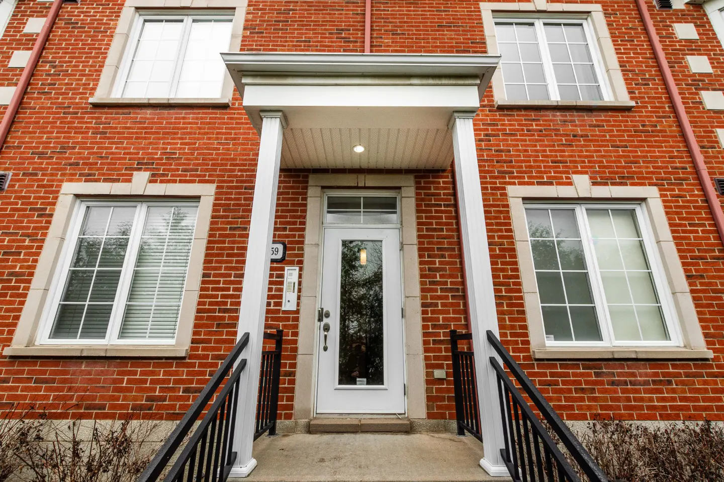 Red brick building with white door and trim. Black railings lead to the entrance. Windows are white with multi-pane design.