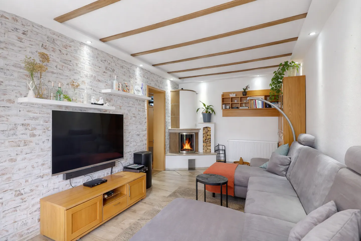 Living room with gray sectional sofa, fireplace, TV, and white brick wall with shelves. Ceiling has wooden beams.